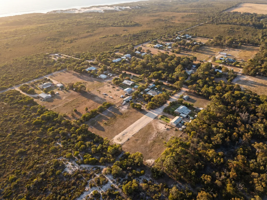 Aerial view of a coastal-fringe rural-residential cluster in south-west WA, properties with cleared firebreaks visible against coastal heath and the Indian Ocean in the distance