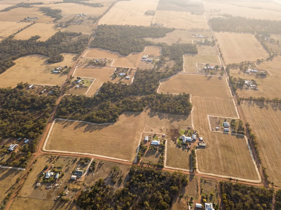 Aerial view of wheatbelt-edge landscape in WA showing scattered rural-residential properties between agricultural paddocks of golden stubble and remnant eucalypt bushland
